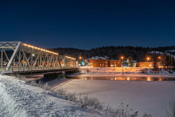 A lit steel truss bridge over a frozen river at night, with low brick buildings, orange street lights and dark spruce hills beyond the snow